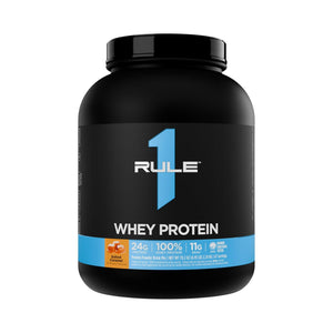 A black tub of RULE 1 Whey Protein in Vanilla Caramel flavor, featuring a black label with a prominent blue number 1 and a blue band at the bottom.